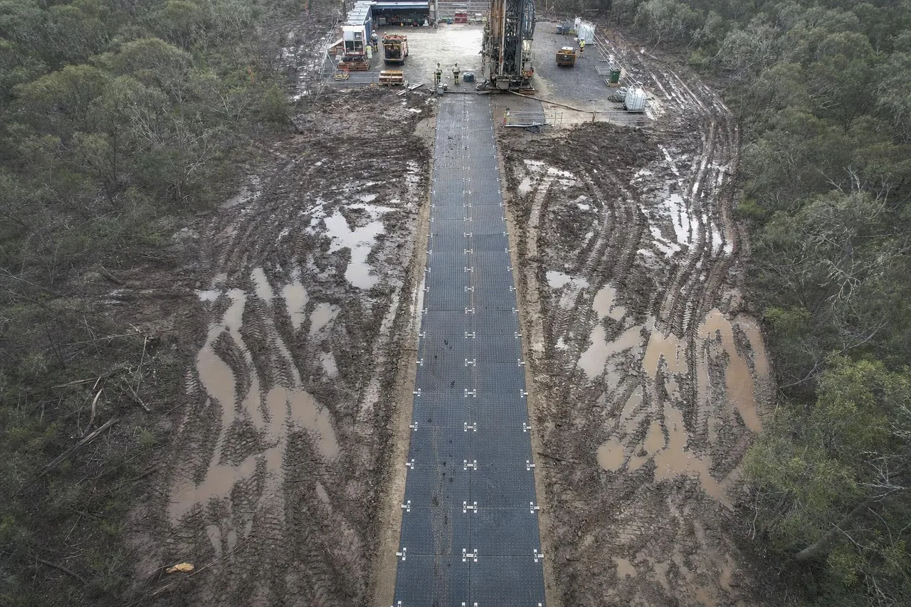 Looking down on a straight road of black bog mats laid across churned mud toward a drilling rig